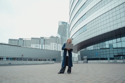Caucasian woman in blue coat working on a laptop outside modern high-rise buildings.
