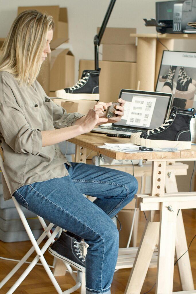 Woman using laptop and smartphone to manage an online shoe store in a workspace filled with boxes.