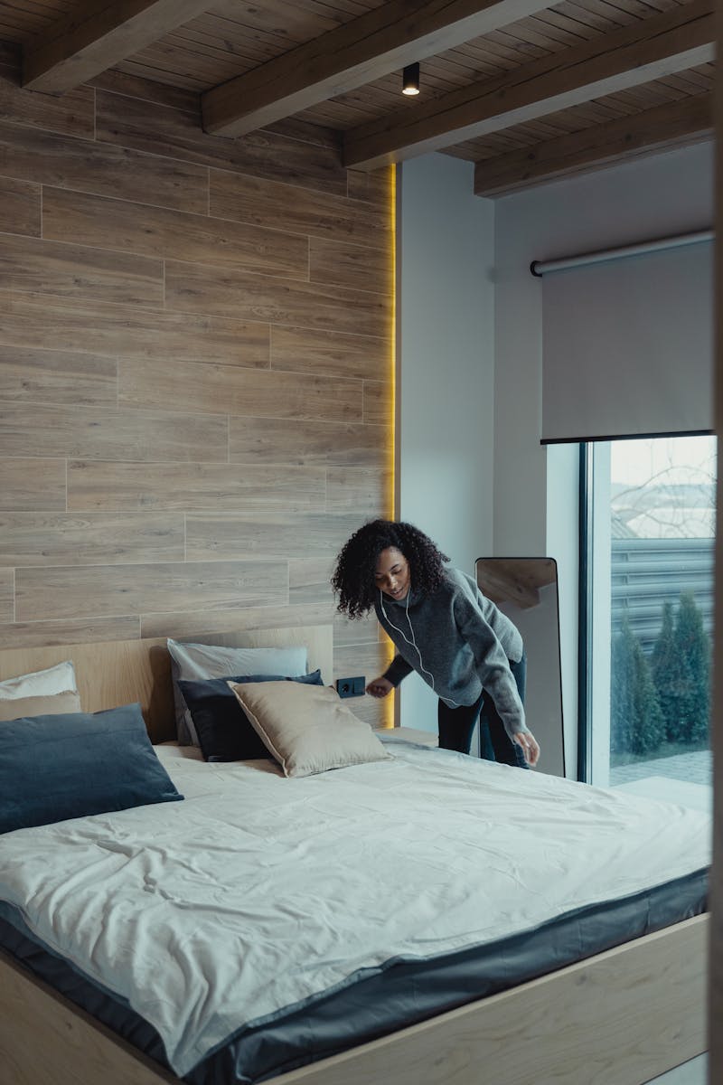African American woman fixing pillows in a modern bedroom with wooden accents, promoting cleanliness and organization.