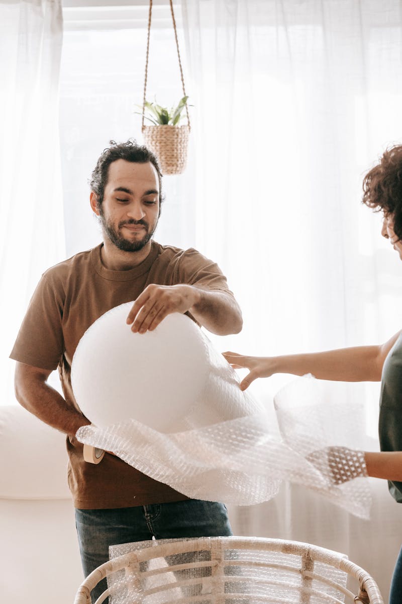 A couple wrapping a chair with bubble wrap in a bright, sunlit room, symbolizing moving or relocating.